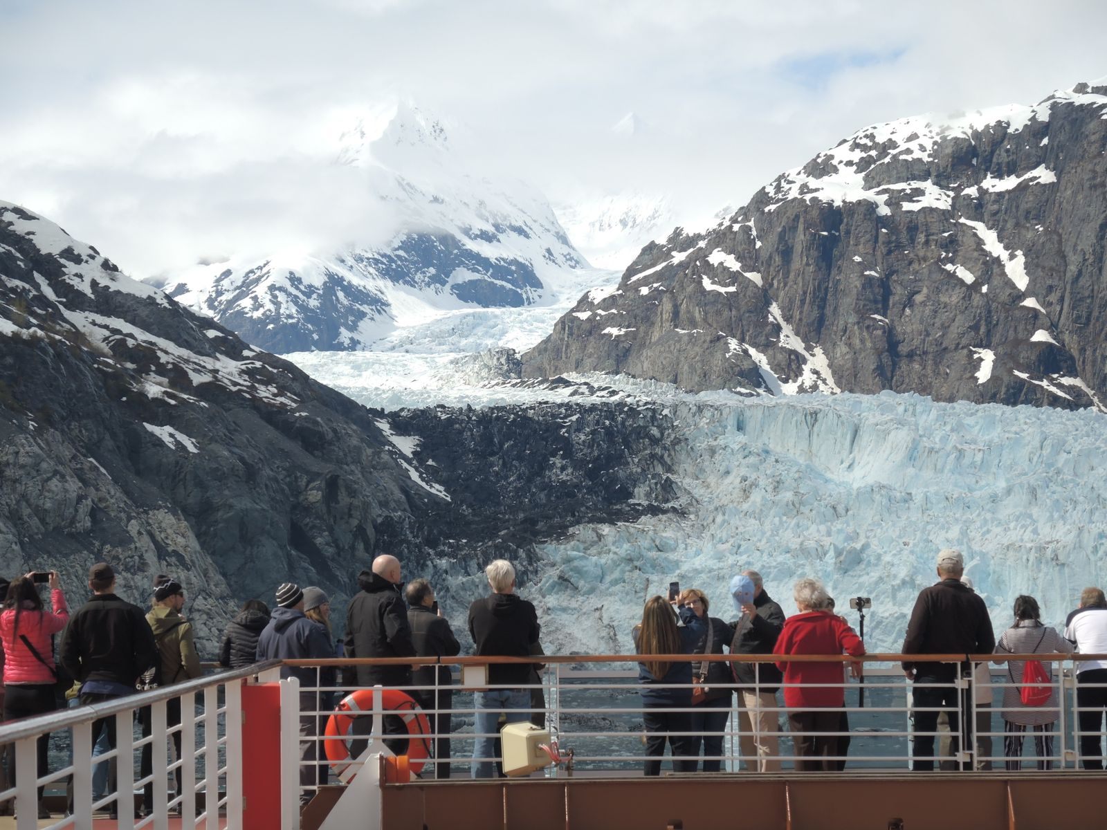 Passengers crowding the deck rail of a ship to watch a tidewater glacier between snowy peaks, the moment that decides which cruise line is best for Alaska for many travelers