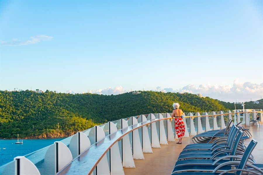 A guest at the rail of a long curved promenade deck lined with loungers, with green hills across the water
